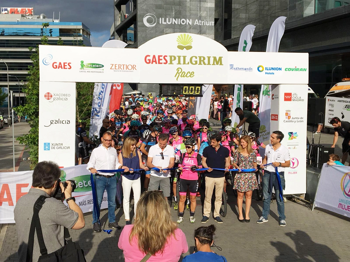 Salida de una carrera ciclista multitudinaria bajo un arco de meta con el nombre “GAES Pilgrim Race”. En primer plano, varias personas sujetan una cinta azul durante el acto inaugural mientras decenas de ciclistas con casco esperan detrás de la línea de salida. El arco incorpora logotipos de patrocinadores e instituciones, y alrededor se observan fotógrafos, banderas promocionales y edificios urbanos en un entorno exterior iluminado por la luz del sol.