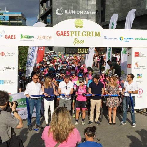 Salida de una carrera ciclista multitudinaria bajo un arco de meta con el nombre “GAES Pilgrim Race”. En primer plano, varias personas sujetan una cinta azul durante el acto inaugural mientras decenas de ciclistas con casco esperan detrás de la línea de salida. El arco incorpora logotipos de patrocinadores e instituciones, y alrededor se observan fotógrafos, banderas promocionales y edificios urbanos en un entorno exterior iluminado por la luz del sol.