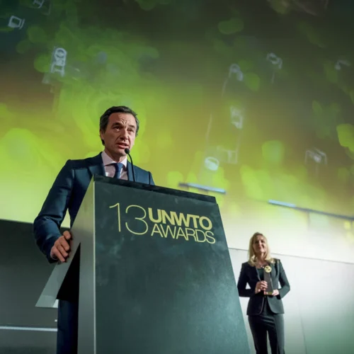 Persona hablando desde un atril durante una ceremonia de premios. En el frontal del atril aparece el texto “13 UNWTO Awards”, mientras otra persona permanece al fondo sosteniendo un galardón. La escena se desarrolla en un auditorio con iluminación verde y una gran pantalla de fondo.