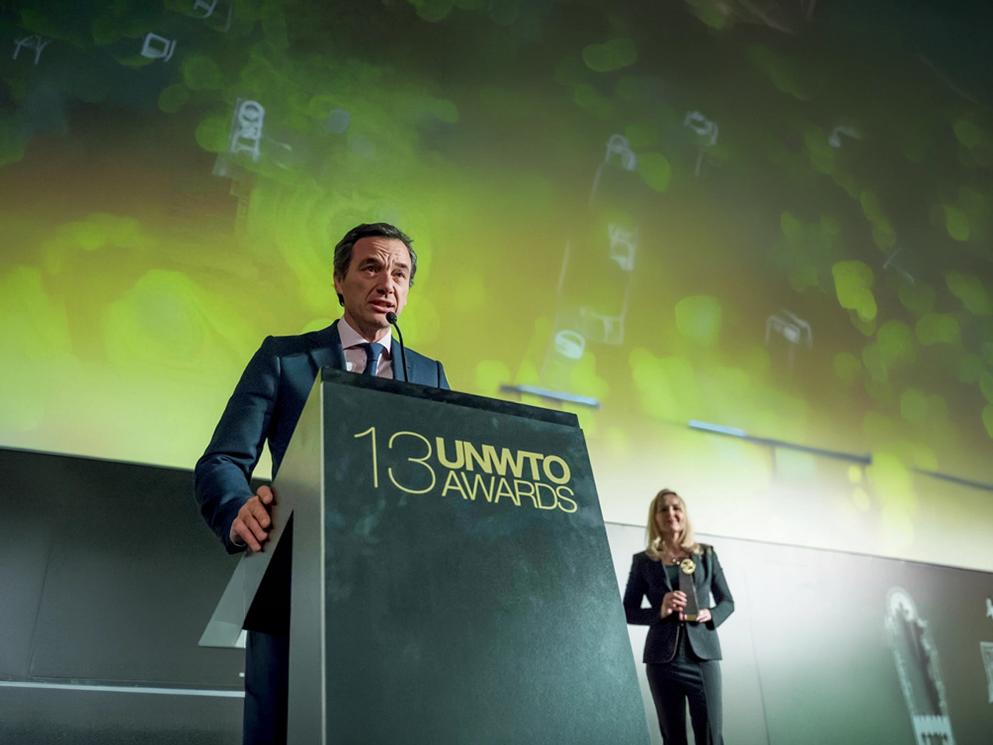 Persona hablando desde un atril durante una ceremonia de premios. En el frontal del atril aparece el texto “13 UNWTO Awards”, mientras otra persona permanece al fondo sosteniendo un galardón. La escena se desarrolla en un auditorio con iluminación verde y una gran pantalla de fondo.