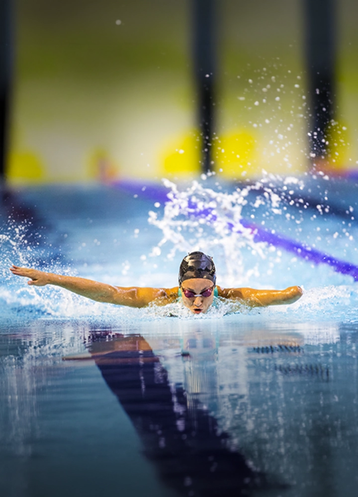 Persona nadando estilo mariposa en una piscina cubierta, captada de frente en pleno movimiento. El agua salpica alrededor mientras los brazos se extienden a ambos lados y las líneas de la calle de nado se reflejan sobre la superficie azul.