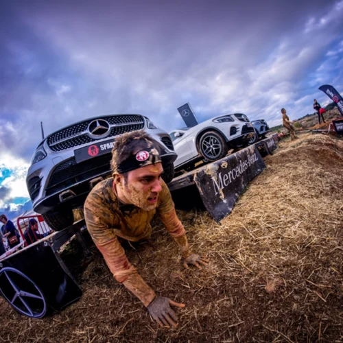 Persona cubierta de barro superando un obstáculo en una carrera al aire libre, arrastrándose bajo una plataforma sobre la que hay varios vehículos Mercedes-Benz. El recorrido discurre sobre paja y tierra, con banderas del evento y un cielo nublado al fondo.