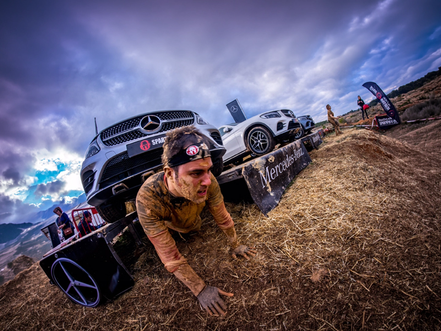 Persona cubierta de barro superando un obstáculo en una carrera al aire libre, arrastrándose bajo una plataforma sobre la que hay varios vehículos Mercedes-Benz. El recorrido discurre sobre paja y tierra, con banderas del evento y un cielo nublado al fondo.