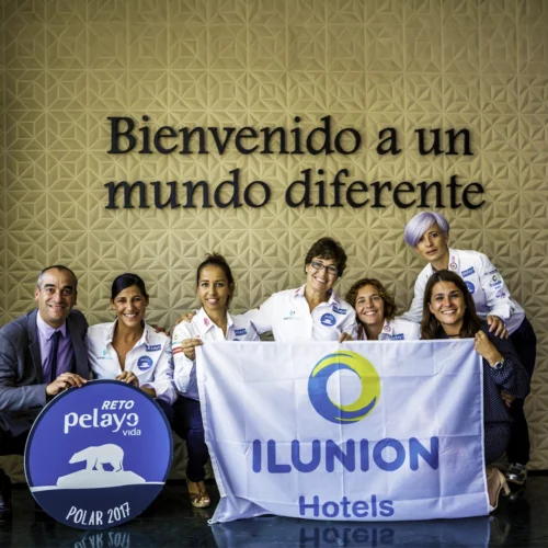 Siete personas posan frente a una pared con el texto “Bienvenido a un mundo diferente”. En primer plano sostienen una bandera de ILUNION Hotels y un cartel circular del “Reto Pelayo Vida Polar 2017”. La fotografía está tomada en un espacio interior con suelo brillante y decoración geométrica en la pared.