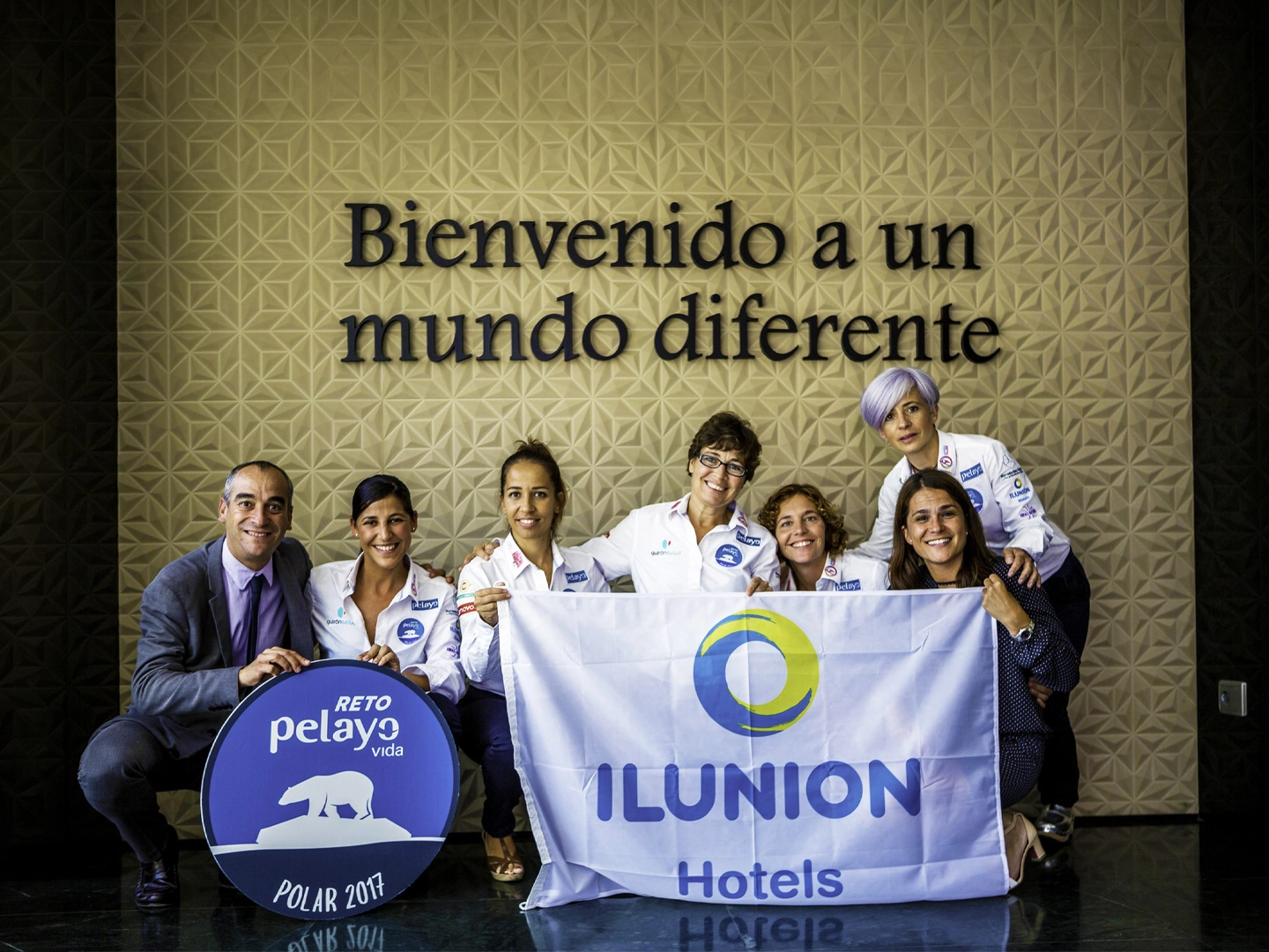 Siete personas posan frente a una pared con el texto “Bienvenido a un mundo diferente”. En primer plano sostienen una bandera de ILUNION Hotels y un cartel circular del “Reto Pelayo Vida Polar 2017”. La fotografía está tomada en un espacio interior con suelo brillante y decoración geométrica en la pared.