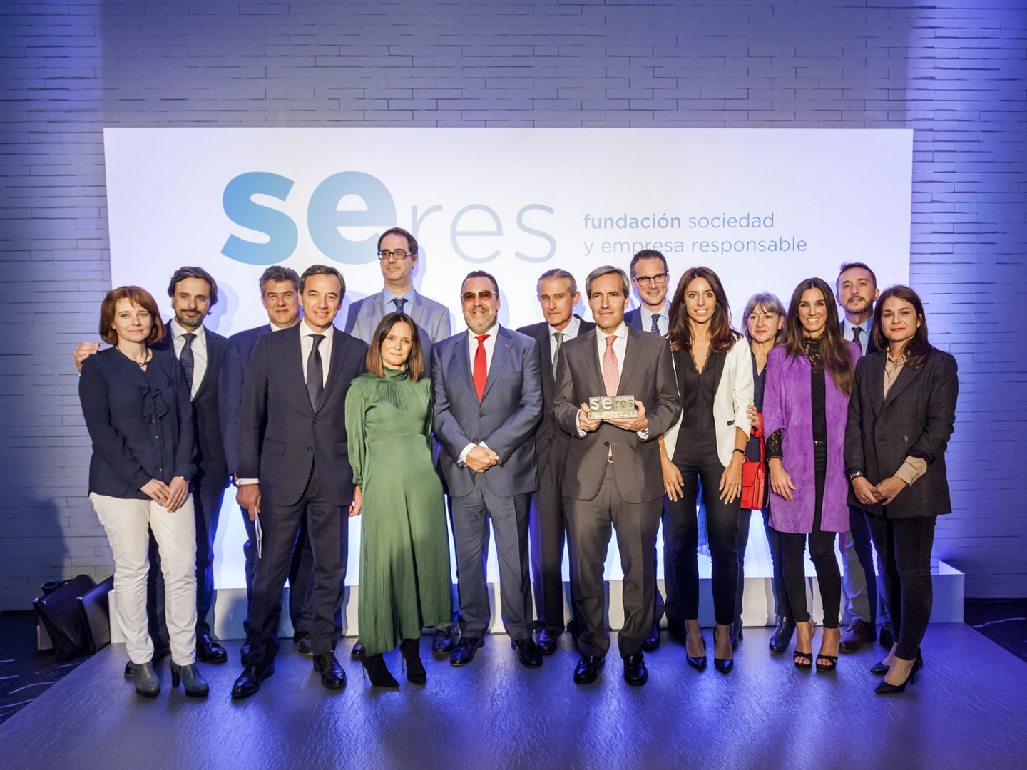 Grupo de personas posando en un escenario durante un acto institucional, delante de un panel con el logotipo de “SERES, fundación sociedad y empresa responsable”. En el centro, una persona sostiene un reconocimiento mientras el resto del grupo se distribuye alrededor. La escena está iluminada con luces azules y blancas.