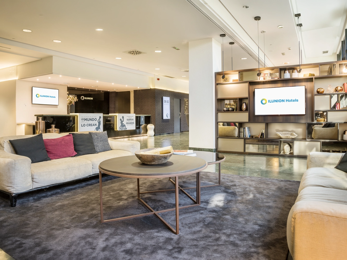 Vestíbulo moderno de un hotel ILUNION con zona de descanso compuesta por sofás beige, cojines decorativos y una mesa redonda sobre una alfombra gris. Al fondo se encuentra la recepción y una estantería con objetos decorativos y una pantalla que muestra el logotipo de ILUNION Hotels. El espacio está iluminado con luz cálida y combina tonos neutros, madera y detalles contemporáneos.