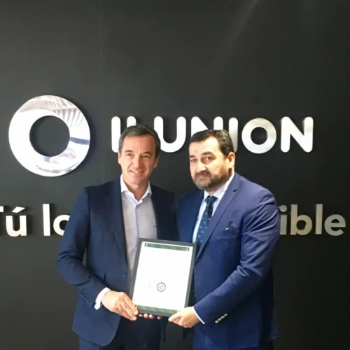 Dos personas con traje posan en el interior de unas oficinas frente a una pared negra con el logotipo de ILUNION y el lema “Tú lo haces posible”. Ambas sostienen un certificado enmarcado en el centro de la imagen. El espacio cuenta con suelo de mármol y asientos bajos oscuros a ambos lados.
