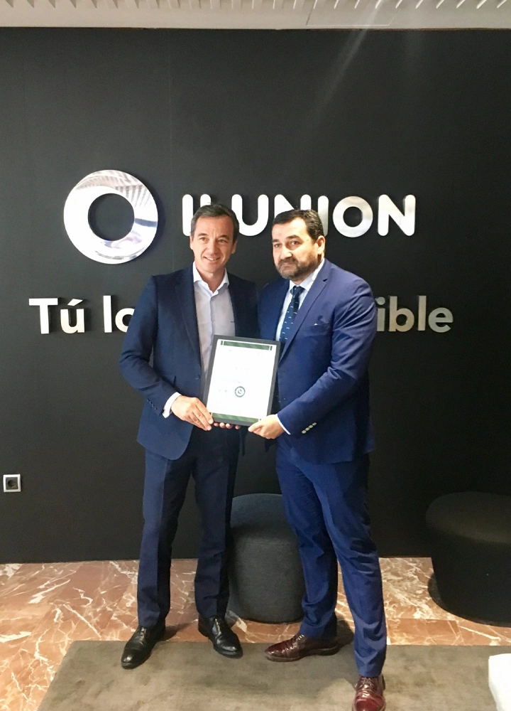 Dos personas con traje posan en el interior de unas oficinas frente a una pared negra con el logotipo de ILUNION y el lema “Tú lo haces posible”. Ambas sostienen un certificado enmarcado en el centro de la imagen. El espacio cuenta con suelo de mármol y asientos bajos oscuros a ambos lados.