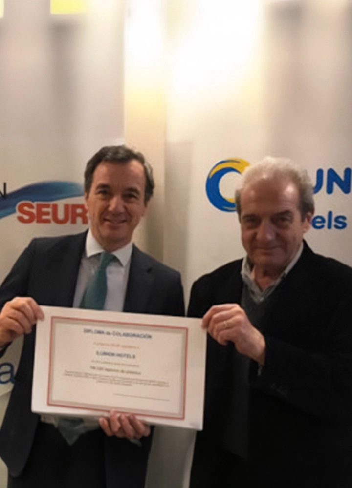 Dos personas sostienen un diploma de reconocimiento durante un acto institucional celebrado en un espacio interior. Detrás aparecen dos paneles corporativos con los logotipos de SEUR e ILUNION Hotels, mientras el certificado ocupa el centro de la escena.