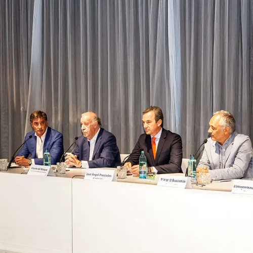 Mesa de conferencia con cuatro ponentes vestidos de manera formal, sentados detrás de un largo escritorio blanco. Cada ponente tiene frente a sí una botella de agua y un cartel con su nombre. El fondo está decorado con cortinas grises que aportan un toque elegante al evento. Uno de los ponentes habla mientras los demás escuchan atentamente.