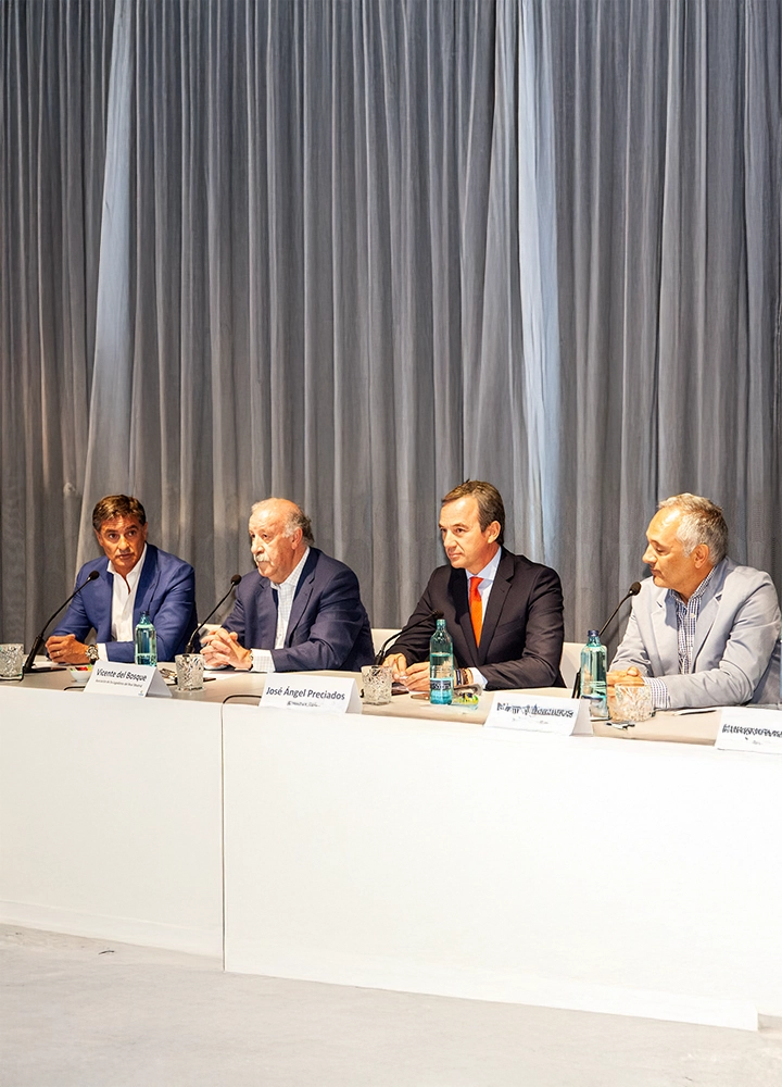 Mesa de conferencia con cuatro ponentes vestidos de manera formal, sentados detrás de un largo escritorio blanco. Cada ponente tiene frente a sí una botella de agua y un cartel con su nombre. El fondo está decorado con cortinas grises que aportan un toque elegante al evento. Uno de los ponentes habla mientras los demás escuchan atentamente.