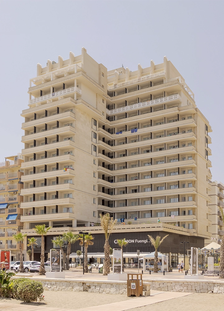 Fachada del hotel ILUNION Fuengirola, un edificio alto de varios pisos con balcones en cada nivel. El hotel está situado frente al paseo marítimo, rodeado de palmeras y vegetación, con un ambiente soleado y claro típico de la costa.