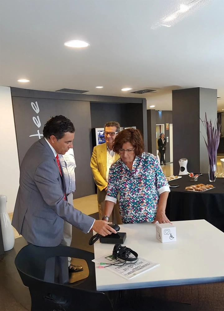 Momento de demostración tecnológica en el lobby de un hotel. Un hombre con traje muestra un dispositivo a una mujer mientras otra persona observa en segundo plano. El entorno es moderno, con decoración minimalista y mesas con aperitivos.