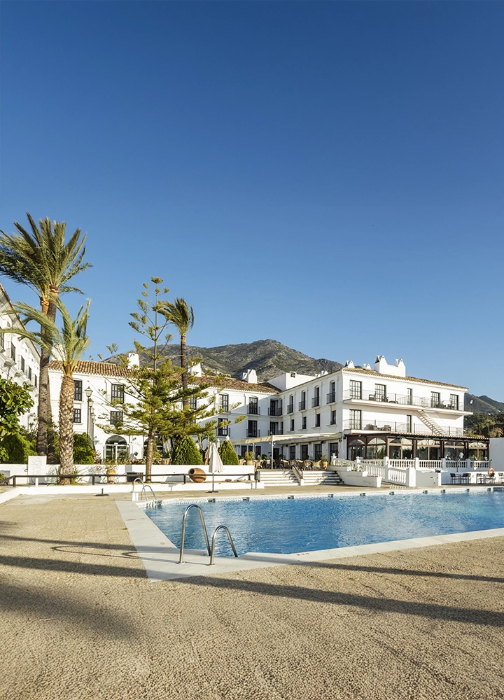 Vista de un hotel de estilo tradicional andaluz con fachadas blancas, palmeras y una piscina al aire libre en primer plano. Al fondo, montañas bajo un cielo azul despejado.