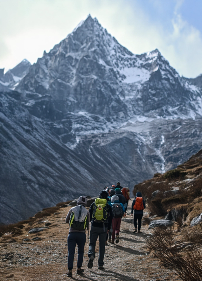 Group of hikers with backpacks walking toward a snowy mountain along a trail surrounded by rocky landscape.