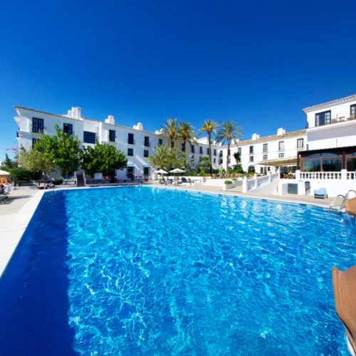 Piscina grande y clara rodeada de edificios blancos con ventanas. Hay palmeras, tumbonas y cielo azul.