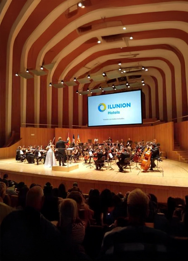 Concierto de orquesta sinfónica en un auditorio con diseño arquitectónico moderno, caracterizado por arcos ondulados en el techo. En el escenario, músicos tocan instrumentos de cuerda y viento, mientras una cantante con vestido claro interpreta frente al director. En la pantalla al fondo, el logotipo de 