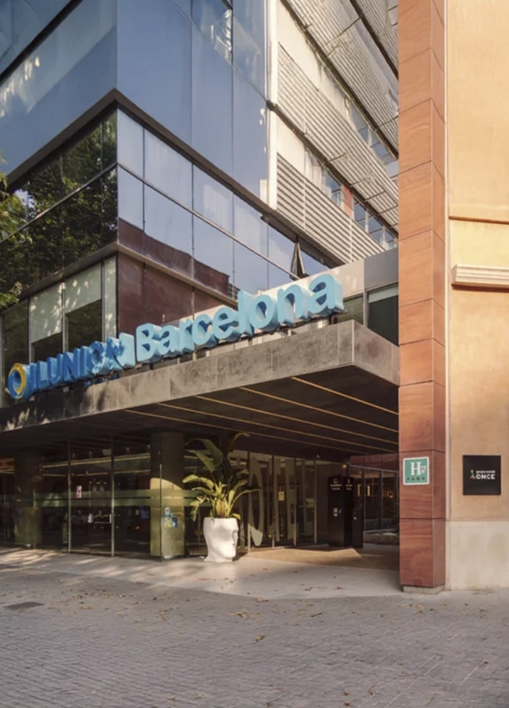 Entrada del edificio ILUNION Barcelona con fachada acristalada y rótulo azul; junto a la puerta, una maceta blanca en forma de cabeza con plantas verdes y señales que indican su categoría hotelera. El diseño moderno y cuidado transmite profesionalidad y hospitalidad.
