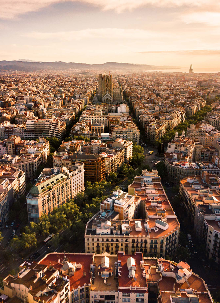 Aerial view of Barcelona during golden hour, with the Sagrada Família standing out among the grid-like layout of the Eixample district. The warm light enhances the architectural beauty of the city and its iconic landmark.