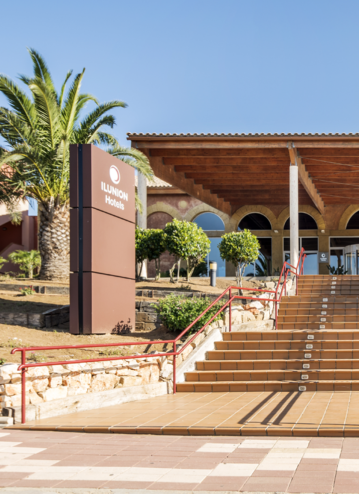 Building entrance with wide stairs, wooden ceiling, and columns. Arched windows, palm trees, 'ILUNION Hotels' sign. Blue sky.