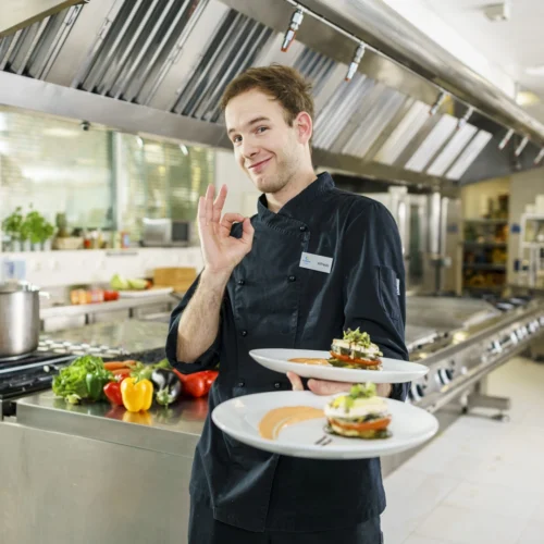 La imagen muestra a una persona vestida con uniforme de cocina oscuro en una cocina profesional de acero inoxidable, sosteniendo dos platos cuidadosamente emplatados. La escena se desarrolla en un entorno gastronómico moderno y equipado, donde se aprecian superficies de trabajo industriales, campanas extractoras y diferentes verduras frescas, como pimientos de varios colores, colocadas sobre una mesa de preparación al fondo. Los platos presentan una elaboración culinaria de apariencia refinada, con una composición ordenada de ingredientes, acompañada de salsas y elementos decorativos. Además, la persona realiza un gesto con la mano que suele utilizarse para indicar aprobación o calidad del resultado culinario. En la chaquetilla se observa una identificación con el nombre “Alfredo”. La fotografía transmite una imagen de profesionalidad, cuidado por la presentación de los alimentos y compromiso con la calidad gastronómica dentro de un entorno de restauración profesional.