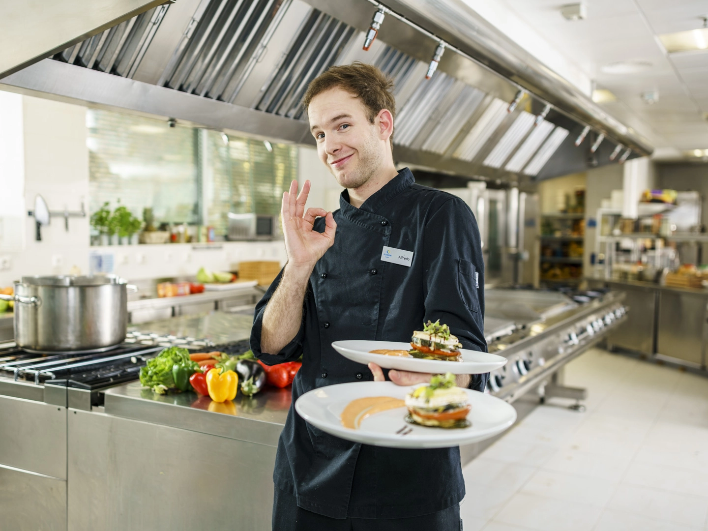 La imagen muestra a una persona vestida con uniforme de cocina oscuro en una cocina profesional de acero inoxidable, sosteniendo dos platos cuidadosamente emplatados. La escena se desarrolla en un entorno gastronómico moderno y equipado, donde se aprecian superficies de trabajo industriales, campanas extractoras y diferentes verduras frescas, como pimientos de varios colores, colocadas sobre una mesa de preparación al fondo. Los platos presentan una elaboración culinaria de apariencia refinada, con una composición ordenada de ingredientes, acompañada de salsas y elementos decorativos. Además, la persona realiza un gesto con la mano que suele utilizarse para indicar aprobación o calidad del resultado culinario. En la chaquetilla se observa una identificación con el nombre “Alfredo”. La fotografía transmite una imagen de profesionalidad, cuidado por la presentación de los alimentos y compromiso con la calidad gastronómica dentro de un entorno de restauración profesional.