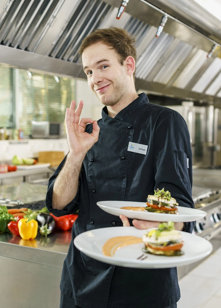 La imagen muestra a una persona vestida con uniforme de cocina oscuro en una cocina profesional de acero inoxidable, sosteniendo dos platos cuidadosamente emplatados. La escena se desarrolla en un entorno gastronómico moderno y equipado, donde se aprecian superficies de trabajo industriales, campanas extractoras y diferentes verduras frescas, como pimientos de varios colores, colocadas sobre una mesa de preparación al fondo. Los platos presentan una elaboración culinaria de apariencia refinada, con una composición ordenada de ingredientes, acompañada de salsas y elementos decorativos. Además, la persona realiza un gesto con la mano que suele utilizarse para indicar aprobación o calidad del resultado culinario. En la chaquetilla se observa una identificación con el nombre “Alfredo”. La fotografía transmite una imagen de profesionalidad, cuidado por la presentación de los alimentos y compromiso con la calidad gastronómica dentro de un entorno de restauración profesional.