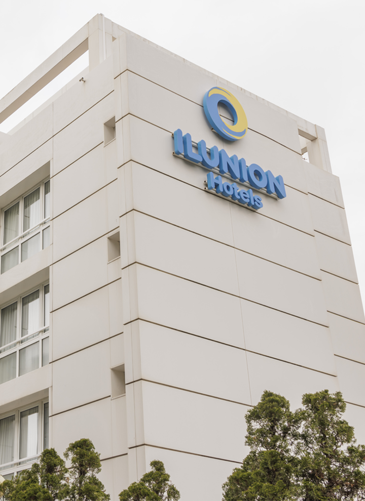 White multi-story building with the 'ILUNION Hotels' logo on the facade. Green trees in front and cloudy sky above.