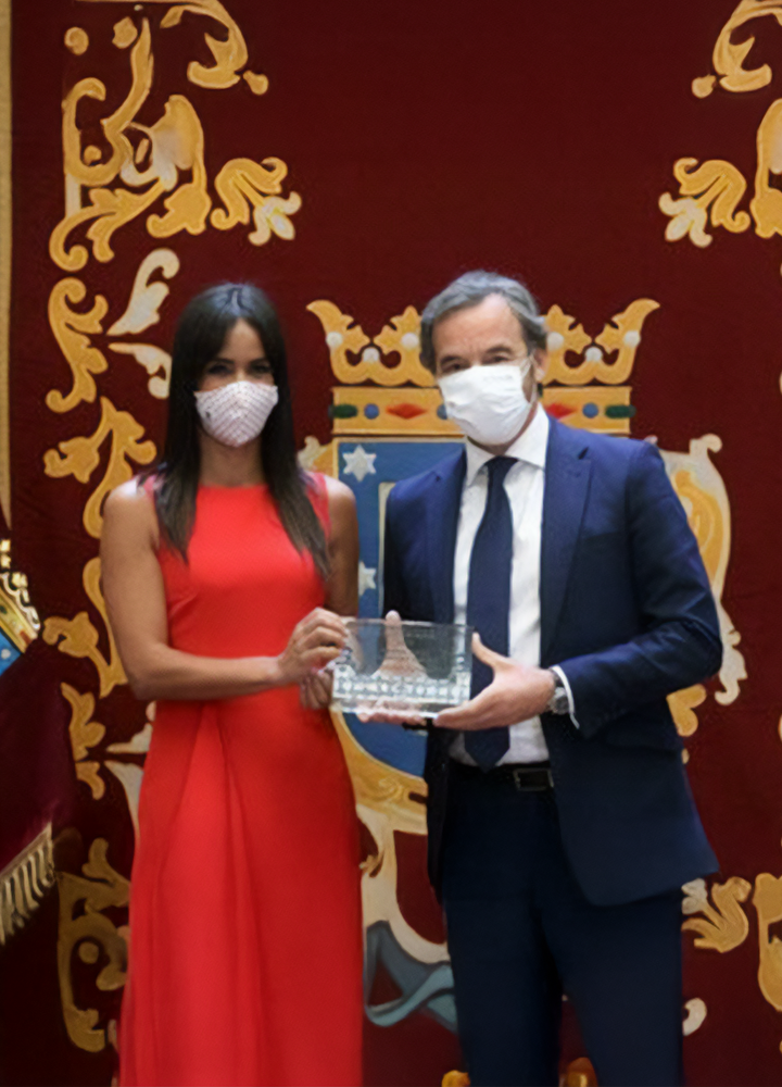 Two people wearing face masks hold an award in front of a decorative backdrop with a coat of arms, during a formal or official ceremony.