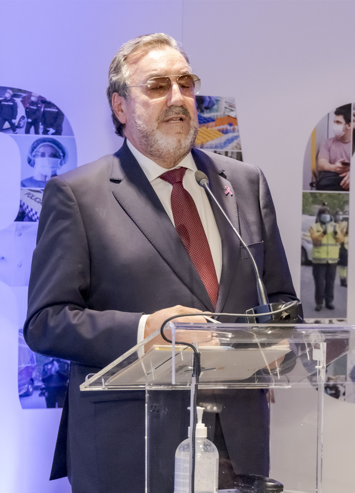 Persona con traje y corbata roja habla en un atril transparente con micrófono; detrás hay imágenes de trabajadores y gel hidroalcohólico sobre el atril.