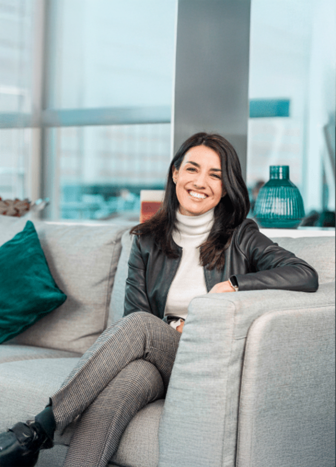 Mujer sentada sonriente en un sofá gris con cojines verdes, vistiendo chaqueta oscura y pantalón de cuadros, en un espacio interior moderno