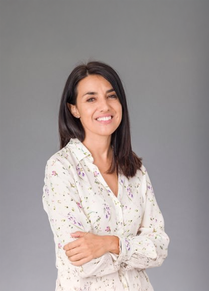 Mujer de pie con camisa clara estampada de flores y brazos cruzados sobre fondo gris liso.