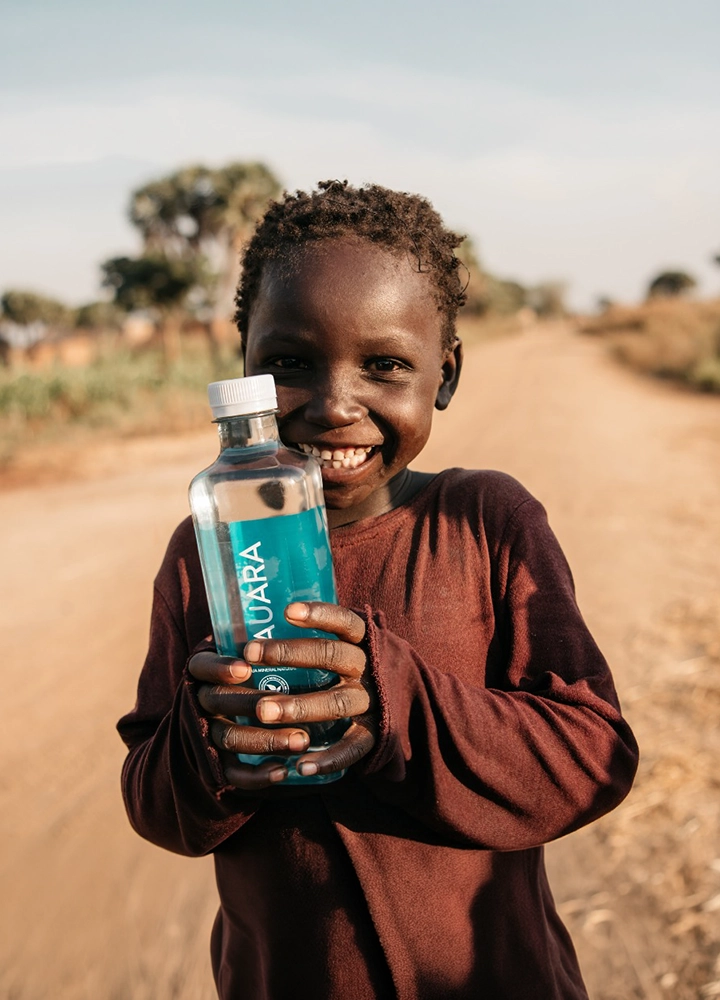 Niño sujetando una botella de agua transparente con etiqueta azul de la marca “AUARA” mientras permanece en un camino de tierra al aire libre. La botella ocupa el primer plano y es sostenida con ambas manos. Al fondo se observan árboles dispersos, vegetación seca y un paisaje rural desenfocado bajo luz natural cálida. La composición centra la atención en la botella y en la persona que la sostiene.