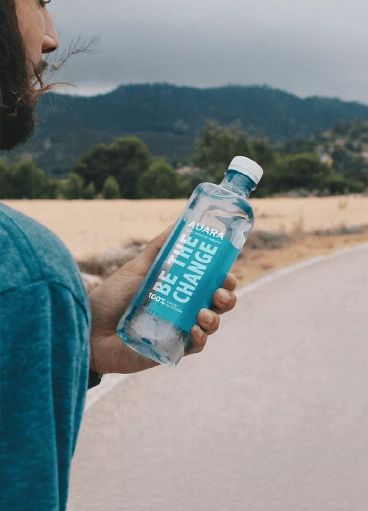 Persona sosteniendo una botella de agua transparente con etiqueta azul en una carretera rural. En la etiqueta se lee “AUARA”, “BE THE CHANGE” y “100% reciclado”. El fondo aparece desenfocado, con campos de tonos dorados, árboles y colinas bajo un cielo grisáceo. La botella ocupa el centro de la imagen y concentra toda la atención, mientras la carretera se extiende hacia la distancia por el lado derecho.