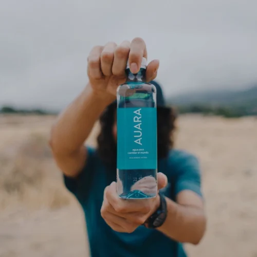 Persona sosteniendo una botella de agua transparente hacia la cámara en un entorno exterior desenfocado de tonos tierra. La etiqueta azul turquesa ocupa el centro de la imagen y contiene el texto “AURA”, junto a las frases “agua para cambiar el mundo” y “agua mineral natural”. La botella aparece enfocada en primer plano, mientras el fondo y la persona quedan difuminados para centrar la atención en el producto.