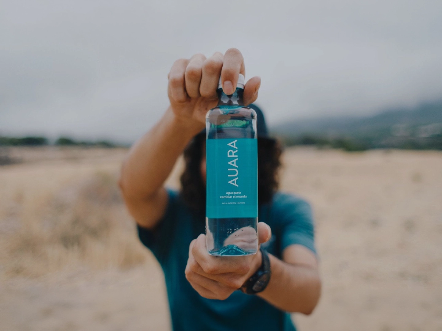 Persona sosteniendo una botella de agua transparente hacia la cámara en un entorno exterior desenfocado de tonos tierra. La etiqueta azul turquesa ocupa el centro de la imagen y contiene el texto “AURA”, junto a las frases “agua para cambiar el mundo” y “agua mineral natural”. La botella aparece enfocada en primer plano, mientras el fondo y la persona quedan difuminados para centrar la atención en el producto.
