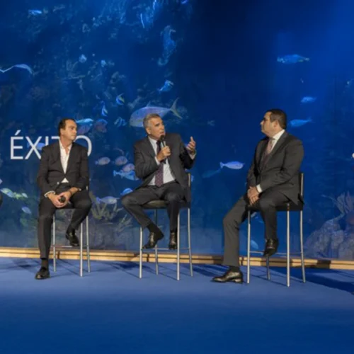 Cinco personas sentadas en sillas altas en un escenario azul, con fondo de acuario y peces visibles, participando en un panel de conversación.