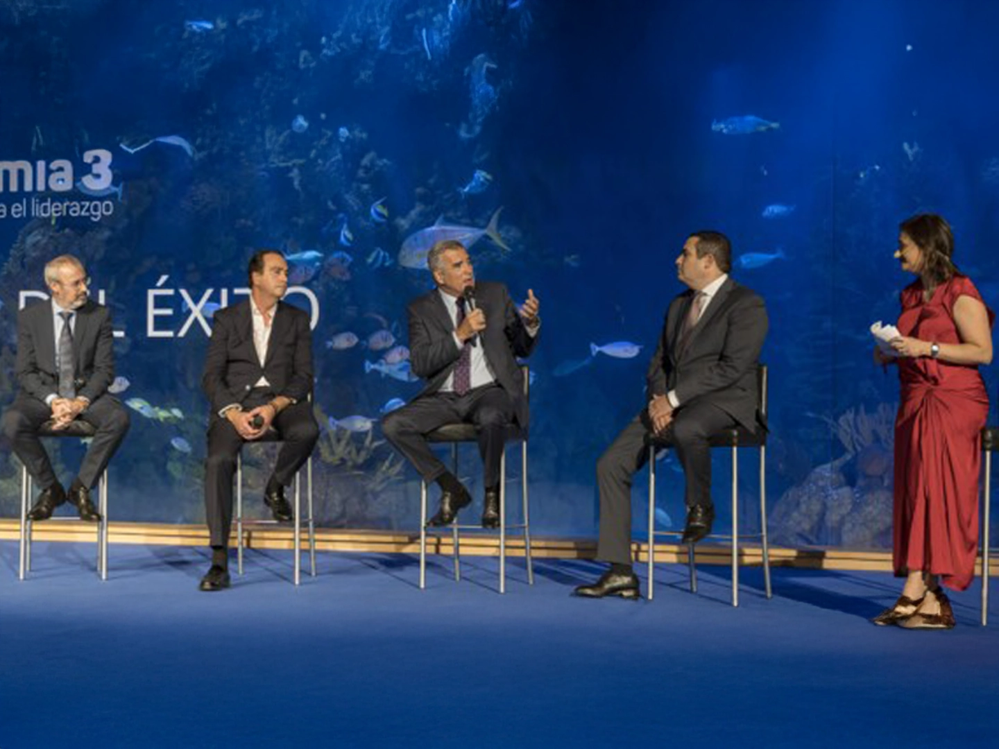 Cinco personas sentadas en sillas altas en un escenario azul, con fondo de acuario y peces visibles, participando en un panel de conversación.