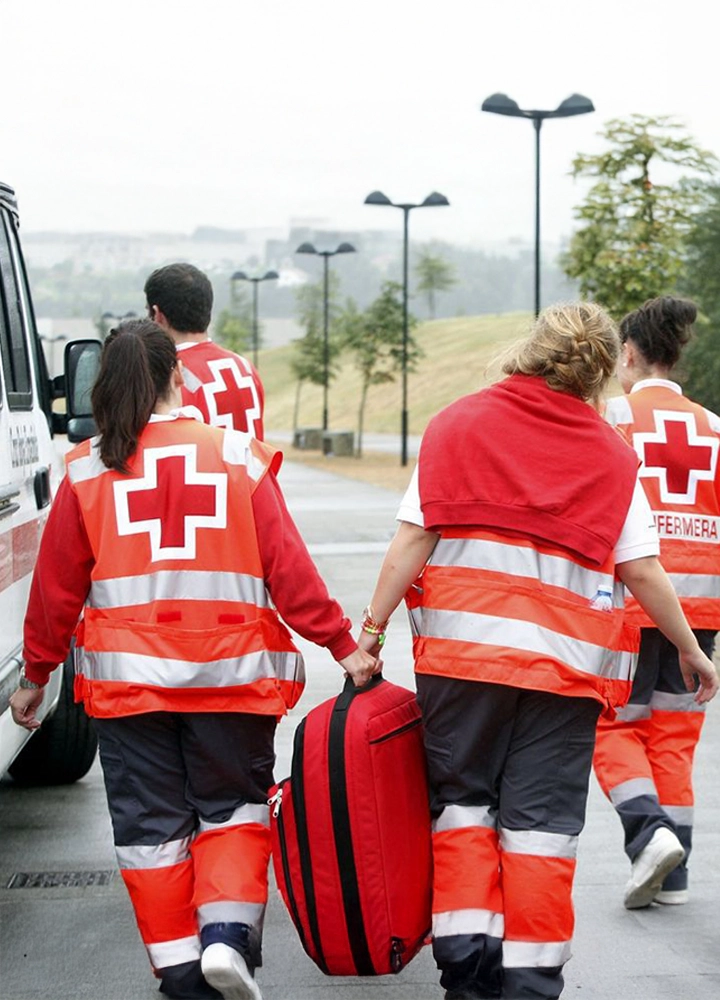 Cuatro personas con chaquetas reflectantes de la Cruz Roja caminan hacia un vehículo; una lleva una maleta roja, listos para una intervención de emergencia.