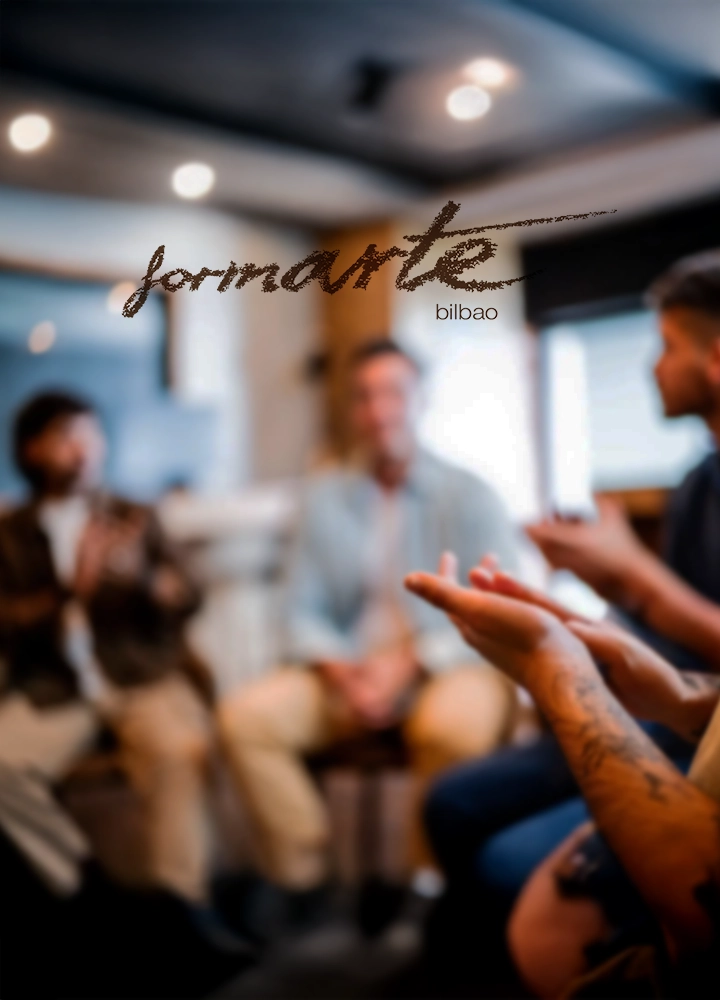 Varias personas se encuentran en una habitación, posiblemente en una reunión o clase. Una persona en primer plano gesticula con las manos. En el centro, el texto 