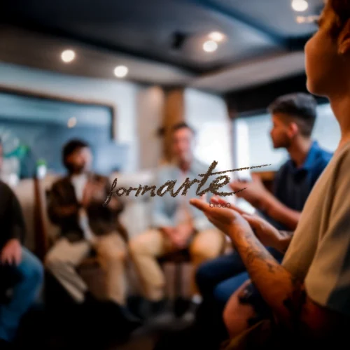 Varias personas se encuentran en una habitación, posiblemente en una reunión o clase. Una persona en primer plano gesticula con las manos. En el centro, el texto 