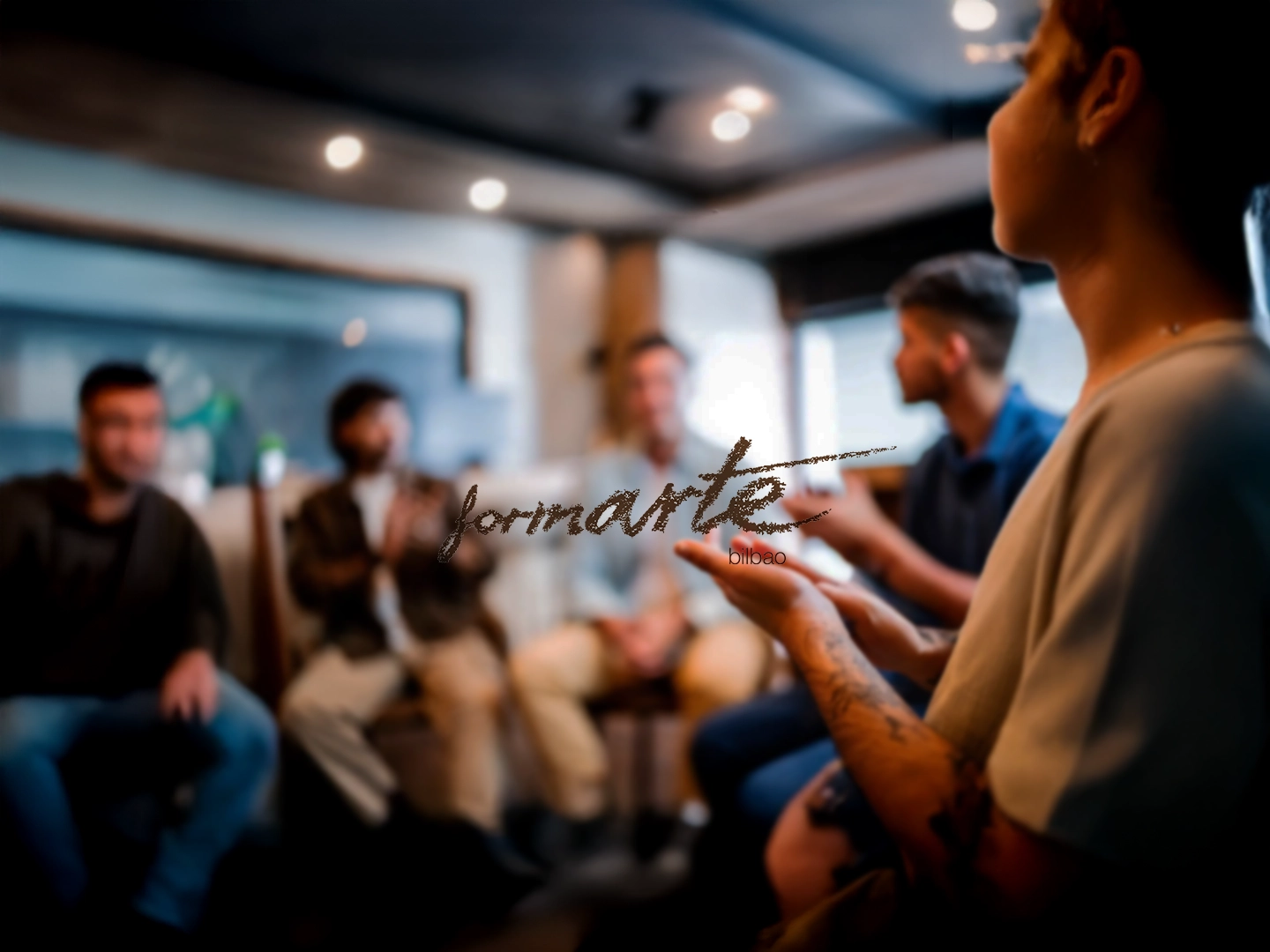Varias personas se encuentran en una habitación, posiblemente en una reunión o clase. Una persona en primer plano gesticula con las manos. En el centro, el texto "formarte bilbao".