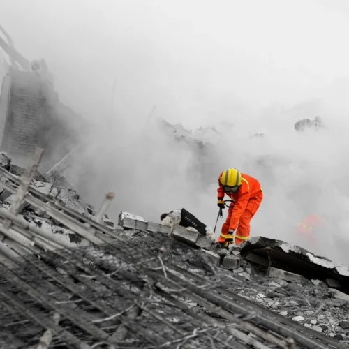 Persona con traje de protección naranja y casco amarillo trabaja sobre un montón de escombros formados por bloques de hormigón rotos y barras metálicas expuestas; el entorno aparece cubierto por una densa nube de polvo o humo gris que reduce la visibilidad, mientras la figura se inclina hacia delante manipulando herramientas entre los restos en una zona de destrucción con estructuras colapsadas en el fondo.