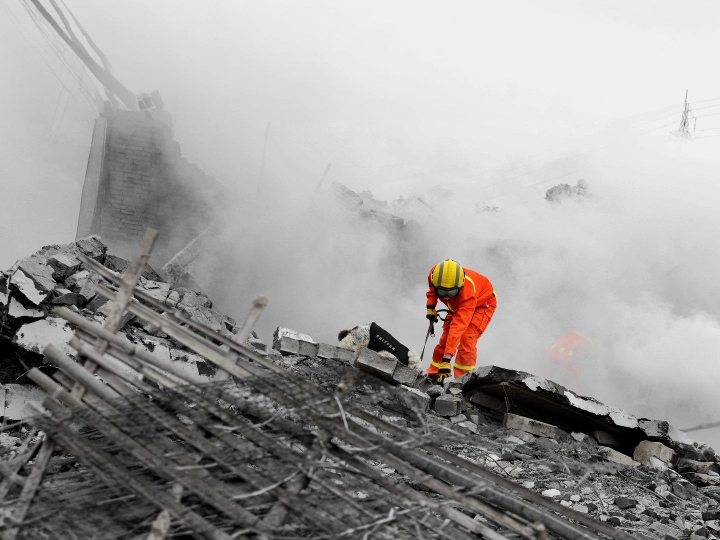 Persona con traje de protección naranja y casco amarillo trabaja sobre un montón de escombros formados por bloques de hormigón rotos y barras metálicas expuestas; el entorno aparece cubierto por una densa nube de polvo o humo gris que reduce la visibilidad, mientras la figura se inclina hacia delante manipulando herramientas entre los restos en una zona de destrucción con estructuras colapsadas en el fondo.