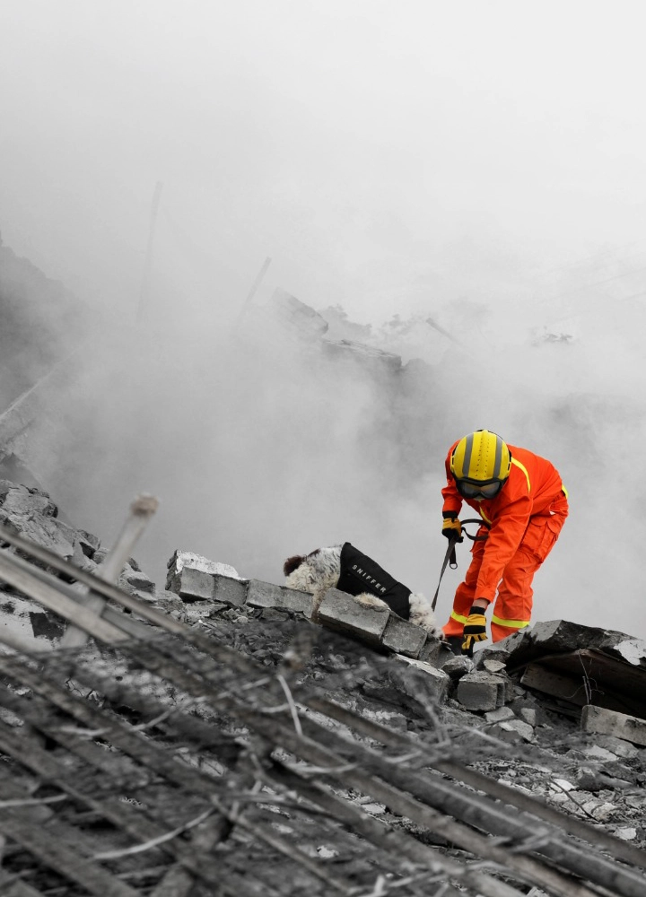 Persona con traje de protección naranja y casco amarillo trabaja sobre un montón de escombros formados por bloques de hormigón rotos y barras metálicas expuestas; el entorno aparece cubierto por una densa nube de polvo o humo gris que reduce la visibilidad, mientras la figura se inclina hacia delante manipulando herramientas entre los restos en una zona de destrucción con estructuras colapsadas en el fondo.