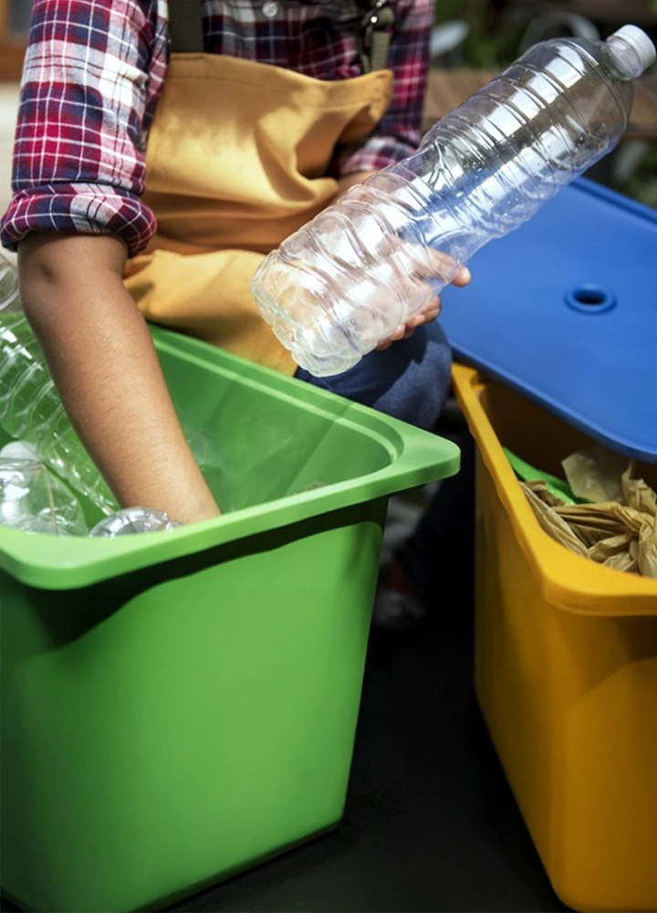Persona con camisa de cuadros y delantal amarillo recicla botellas plásticas en un contenedor verde; al lado, otro contenedor con papel y bolsas.