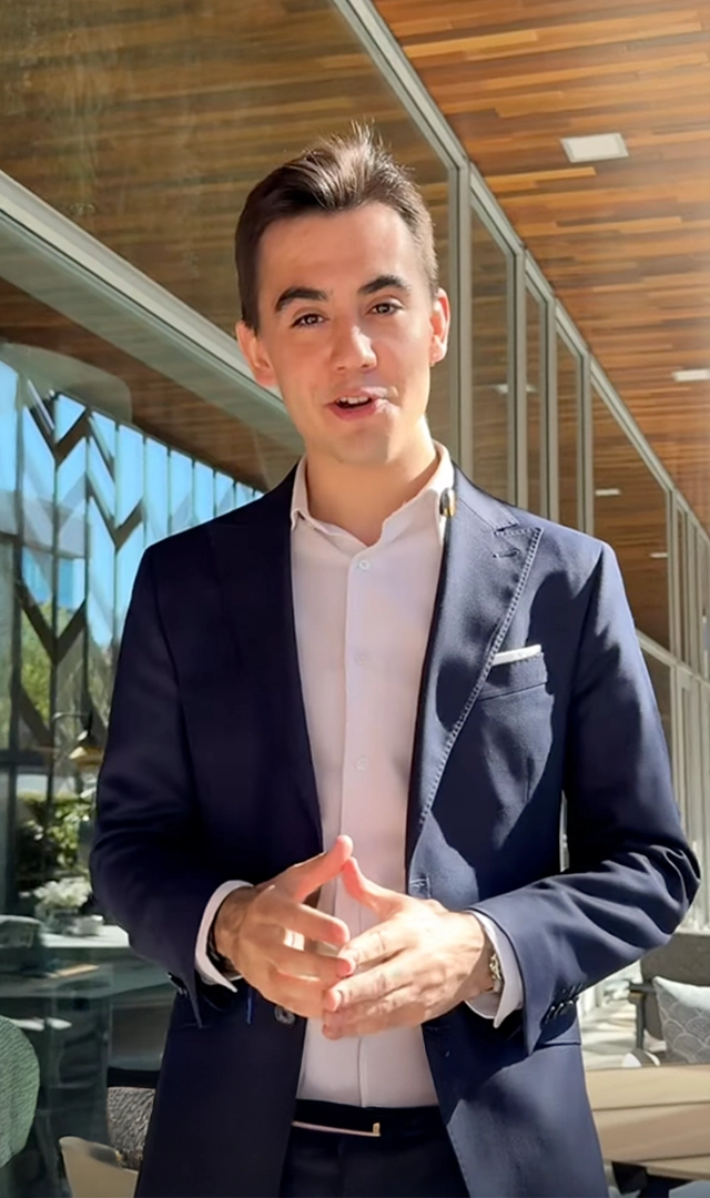 Un hombre joven, vestido con un traje azul marino y una camisa blanca, mira directamente a la cámara mientras junta sus manos. Se encuentra al aire libre con un edificio moderno detrás.