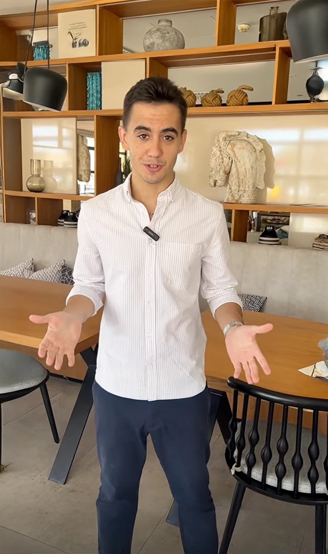 Un hombre joven con una camisa de rayas se para en una habitación con muebles de madera, mirando directamente a la cámara con las palmas de las manos hacia arriba.