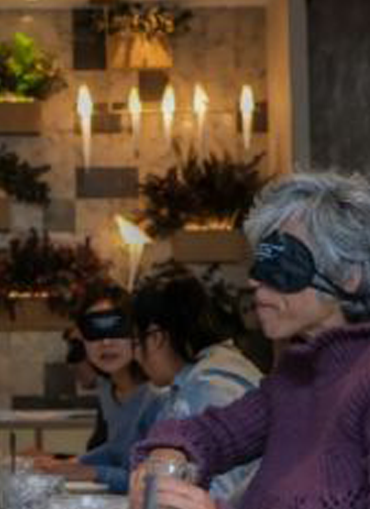 Grupo de personas con los ojos vendados sentados en una mesa durante una actividad sensorial en un entorno decorado con plantas y velas.